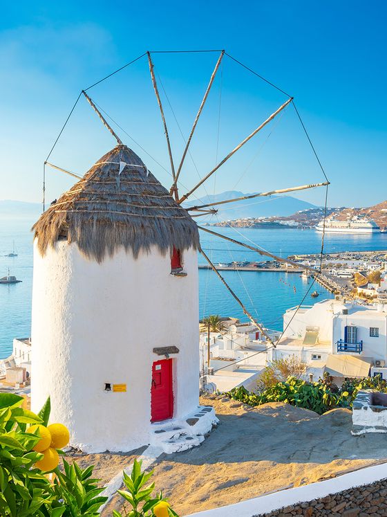 A traditional windmill with a thatched roof and red door overlooks a coastal town and blue sea in Mykonos, Greece.