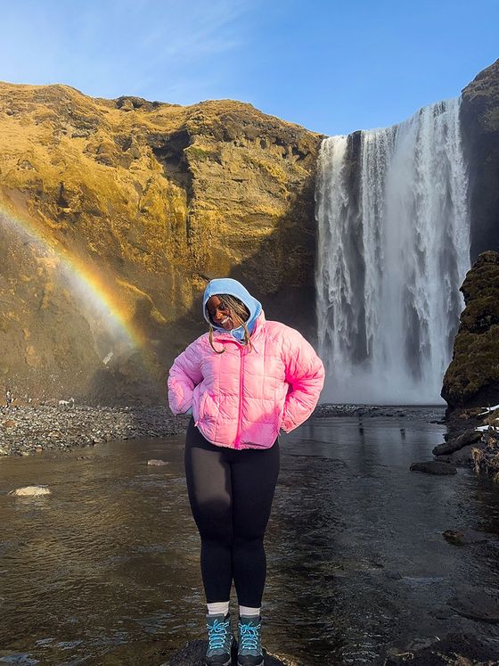 A person in a pink jacket stands by a waterfall and rainbow, against a rocky landscape, under a clear blue sky.