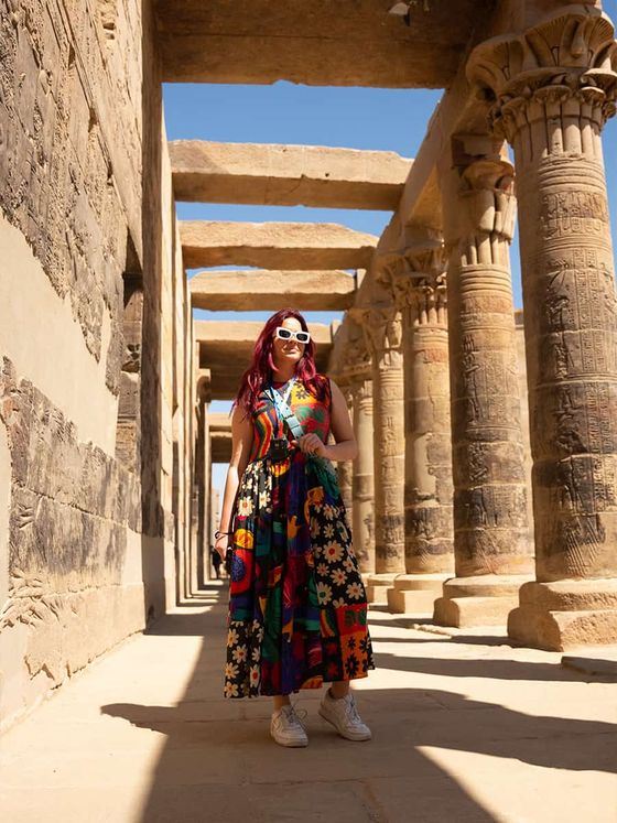 A woman in a colorful dress and sunglasses stands in an ancient stone corridor with engraved columns and bright sunlight.