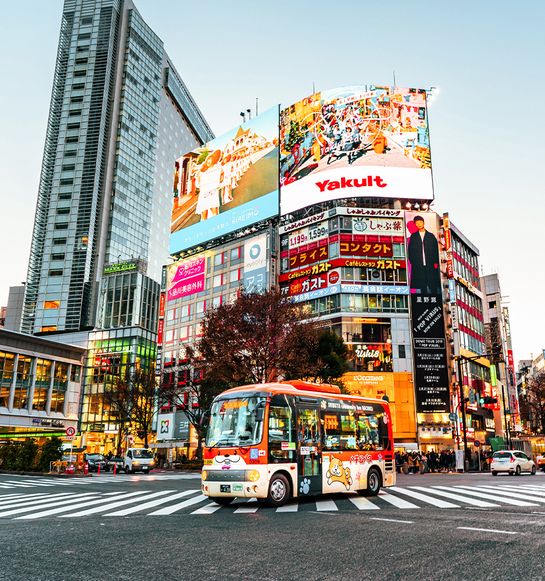A vibrant city intersection with a colorful bus, large digital billboards, and tall buildings during daylight.
