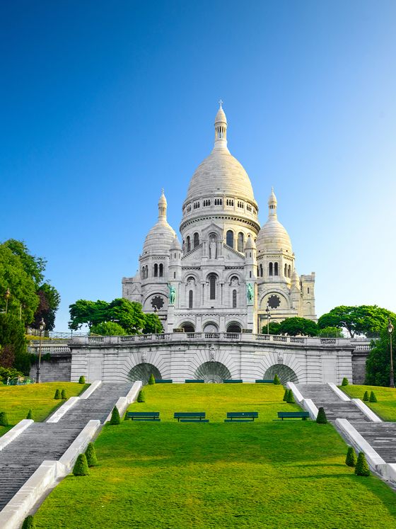 The Sacré-Cœur Basilica in Paris with its white domes, set against a clear blue sky, surrounded by lush greenery and steps leading up to it.