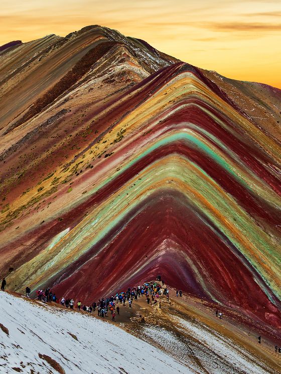 Scenic view of Vinicunca, the Rainbow Mountain in Peru, with vibrant, multicolored layers and a crowd of tourists under a golden sky.