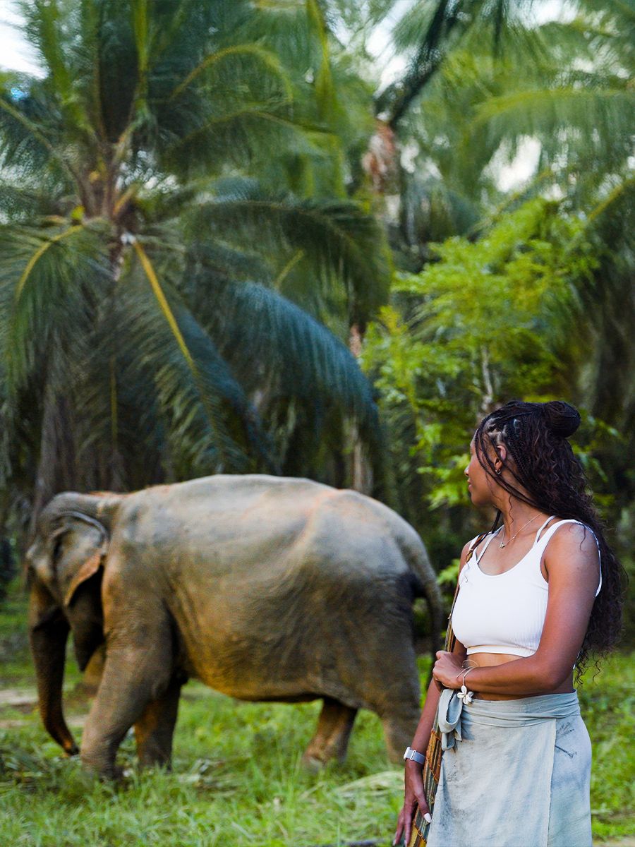 Woman in a white top observing an elephant walking past in a lush, green tropical setting.