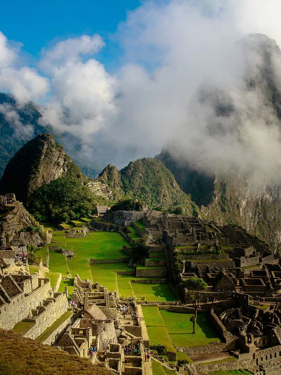 Aerial view of Machu Picchu, showcasing ancient stone ruins, terraced fields, and surrounding misty mountains under a blue sky.