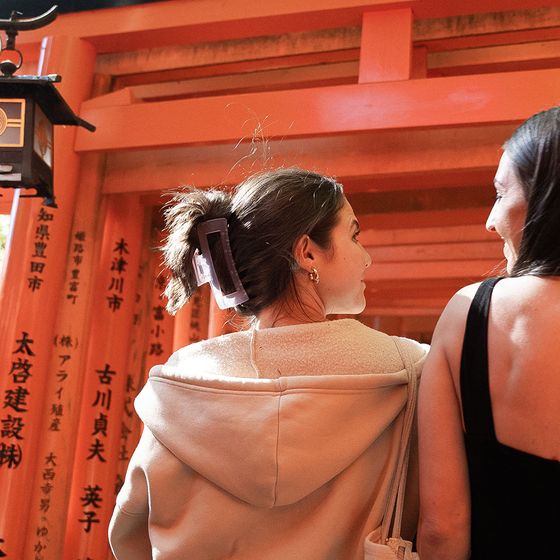 Two people walk through a pathway of vibrant orange torii gates with Japanese inscriptions. One wears a hoodie, the other a sleeveless top.