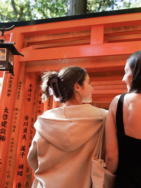 Two people walk through a pathway of vibrant orange torii gates with Japanese inscriptions. One wears a hoodie, the other a sleeveless top.