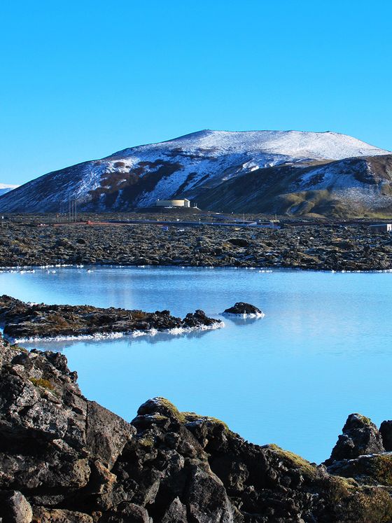 Snow-capped mountain behind a rocky shoreline and serene blue water under a clear sky.