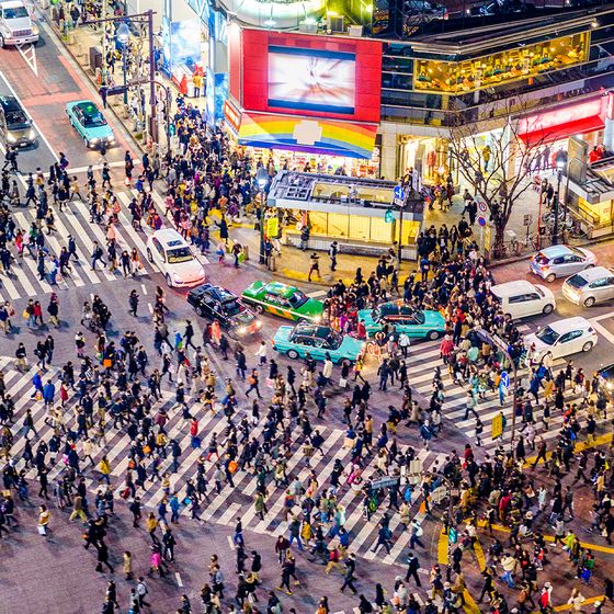 Aerial view of a bustling city intersection at night, crowded with people and cars, illuminated by bright lights and advertisements.