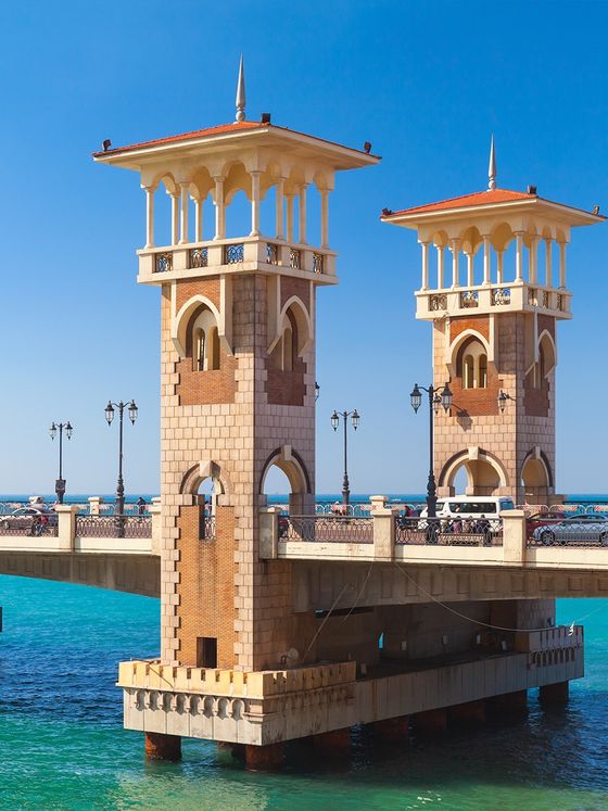Twin ornate towers on a stone bridge over turquoise water, with a clear blue sky in the background.