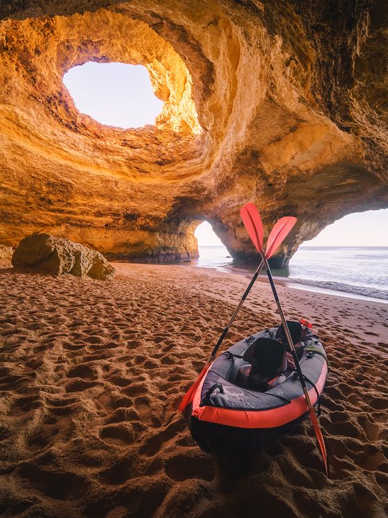 Inflatable kayak with paddles on a sandy beach inside a cave with natural archways and an opening above, revealing a clear sky.