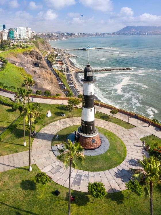 Aerial view of a black and white striped lighthouse surrounded by a park, with a coastline and cityscape in the background under a blue sky.