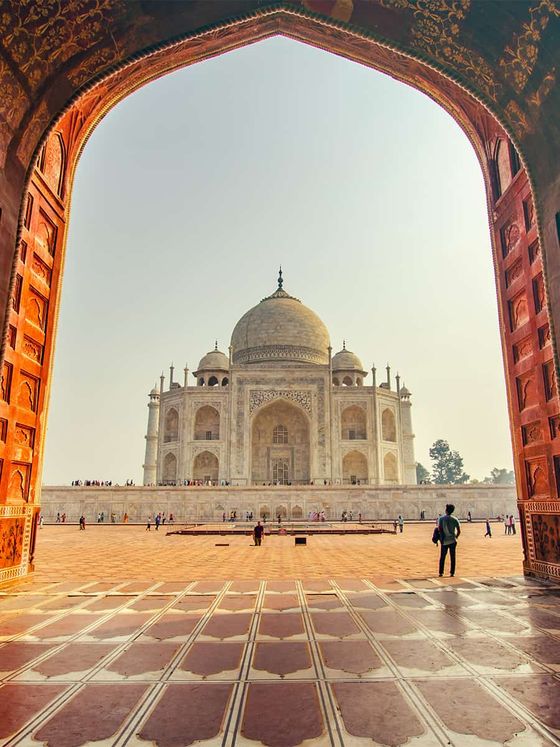 View of the Taj Mahal framed by an ornate archway, with a few visitors walking on the tiled courtyard under a clear sky.