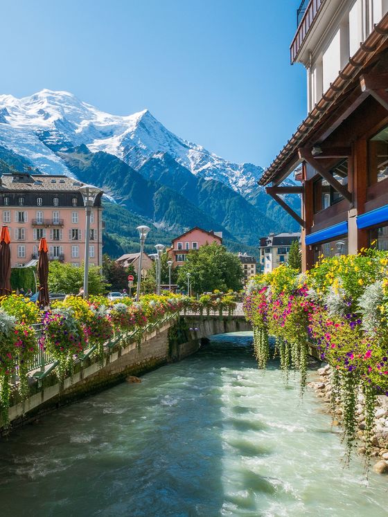 Scenic view of a river lined with vibrant flowers and buildings, with snow-capped mountains under a clear blue sky in the background.