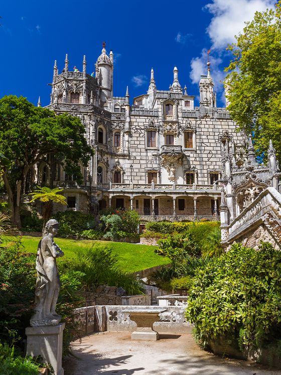 Ornate stone mansion with detailed architecture surrounded by lush greenery, a stone statue, and a garden path under a bright blue sky.