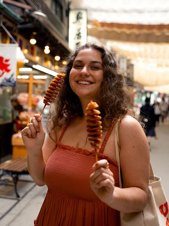 Woman in a rust-colored dress smiles, holding two spiral skewers in a busy indoor market.