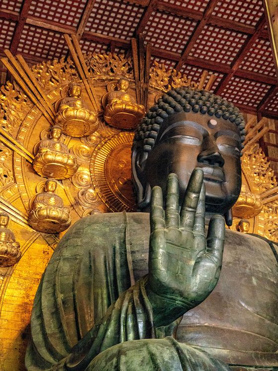 Bronze Buddha statue with an ornate gold halo and raised hand, inside a temple with a patterned ceiling in the background.