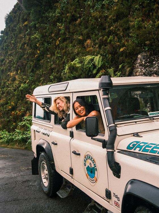 Two people smiling and leaning out of a white SUV with "Green" text, parked on a wet road beside a moss-covered rocky hillside.