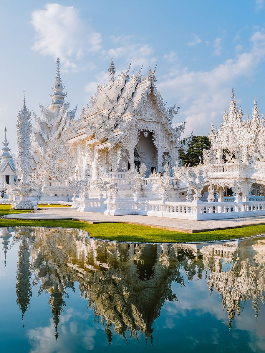 Intricately designed white temple with ornate carvings, reflected in a pond, against a clear blue sky background.