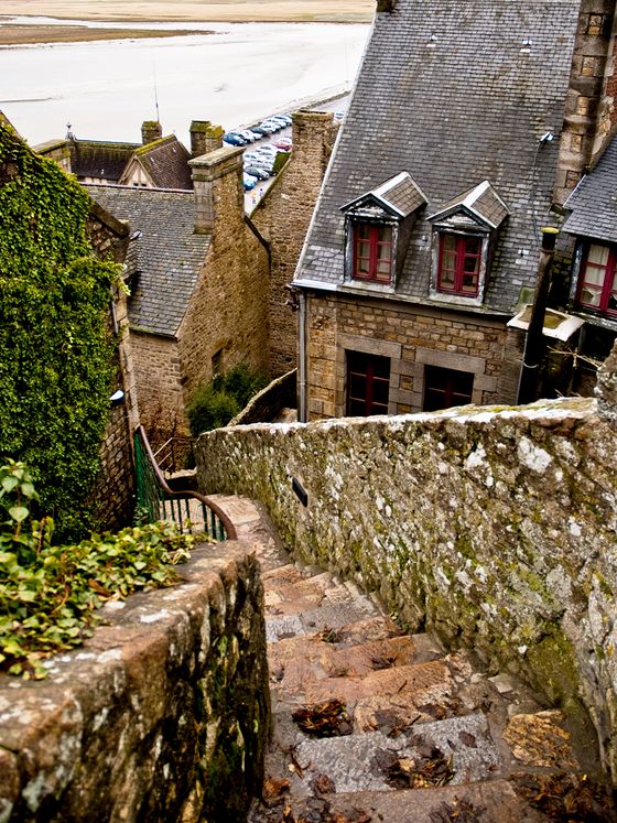 Narrow stone steps descend between ivy-covered medieval buildings with slate roofs, leading toward a distant sandy beach and parked cars.