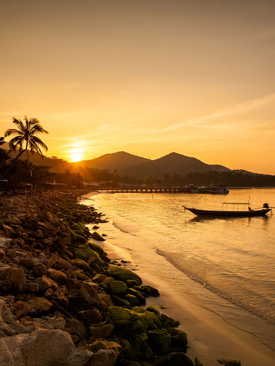 Sunset over a serene beach with a small boat on the calm water, rocky shoreline, palm trees, and silhouetted hills in the background.