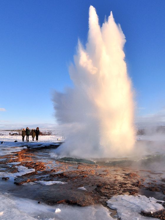 A geyser erupting with a tall burst of water and steam against a clear blue sky, with people observing in the snowy landscape.