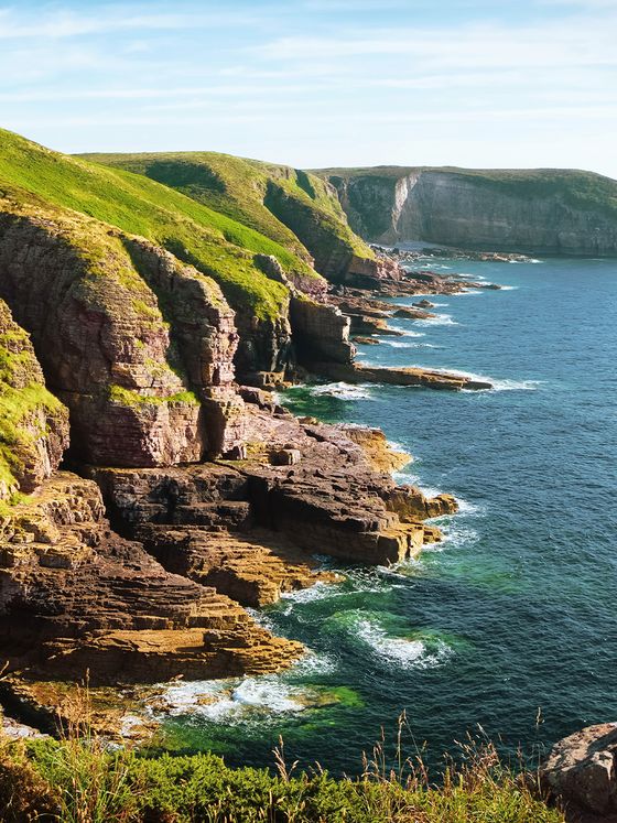 Rocky coastline with green cliffs meeting the blue ocean under a clear sky. Waves gently crash against the rocks in a picturesque scene.