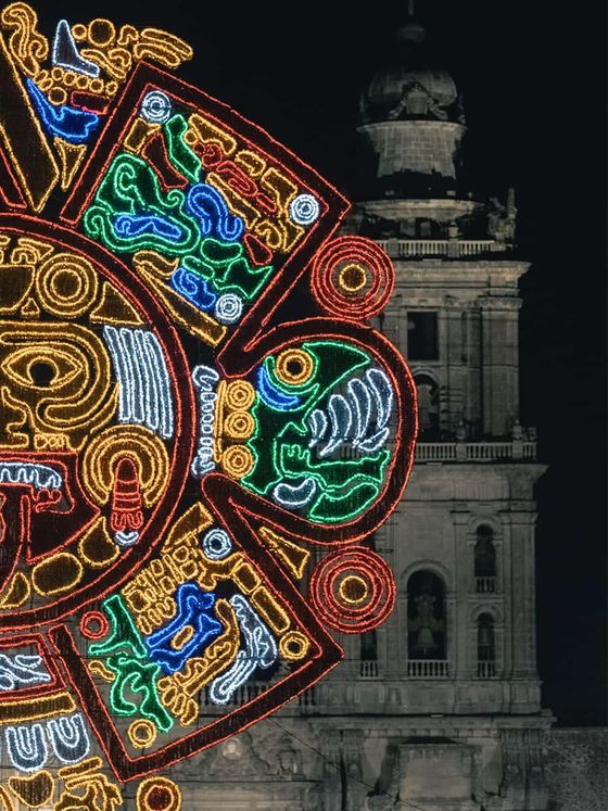 Illuminated Aztec calendar design in vibrant colors beside a historic stone tower at night.