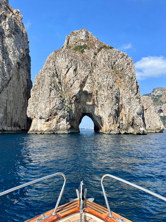 Boat approaching a rugged, towering rock formation with a natural arch over a calm blue sea under a clear sky.