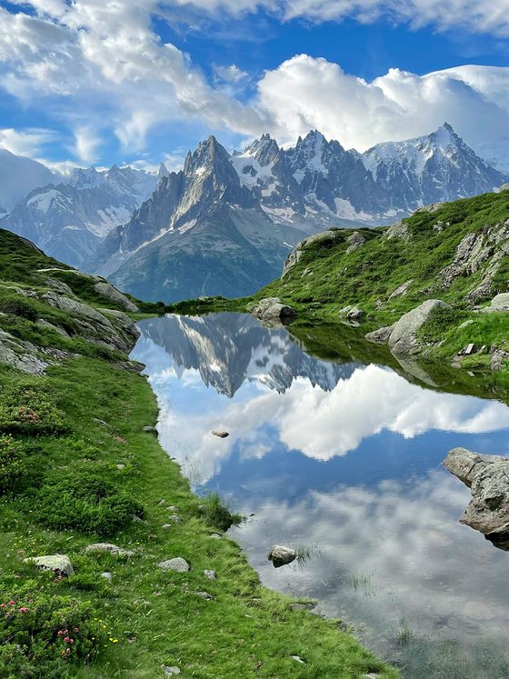 A serene mountain landscape with sharp peaks reflected in a clear, calm lake surrounded by lush green grass and rocks under a partly cloudy sky.