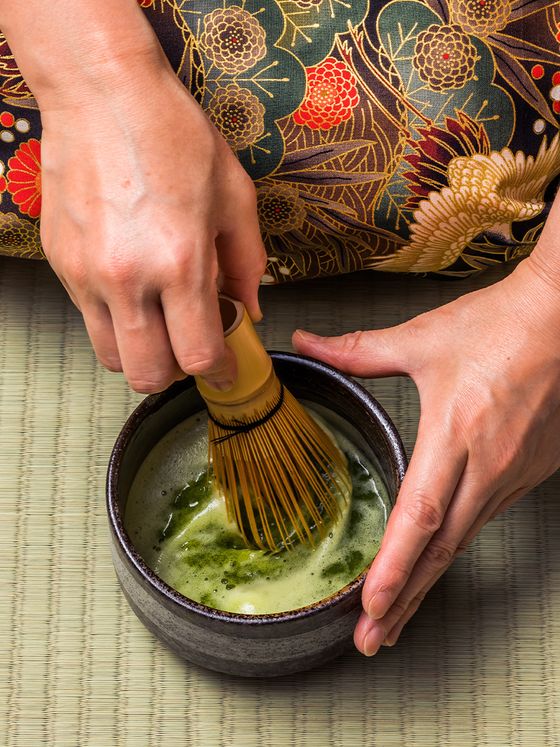 Hands whisking matcha in a bowl on a tatami mat, with a floral-patterned kimono in the background.