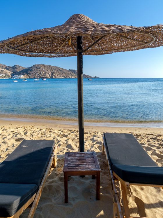 Two lounge chairs under a straw umbrella on a sandy beach overlooking a calm sea with distant mountains on a clear, sunny day.