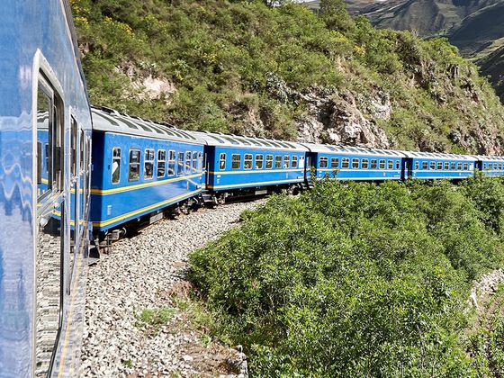 A blue train with multiple cars curves along a lush, green mountainside under a clear blue sky.