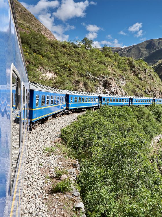 A blue train with multiple cars curves along a lush, green mountainside under a clear blue sky.