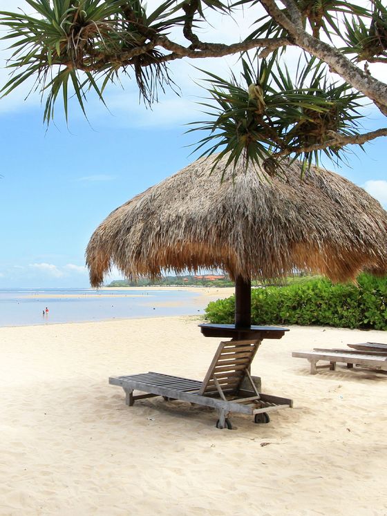 Beach scene with a thatched umbrella, wooden lounge chair, palm trees, and a clear blue sky over a sandy shoreline.