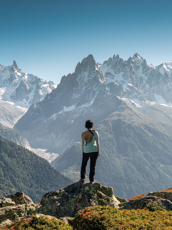A person stands on a rocky ledge, overlooking a range of snow-capped mountains under a clear blue sky.