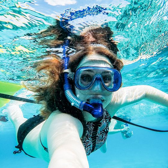 A person snorkels underwater in clear blue water, wearing a snorkel mask and waving at the camera.