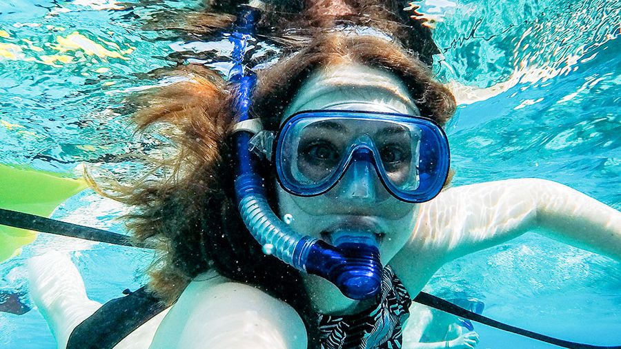 A person snorkels underwater in clear blue water, wearing a snorkel mask and waving at the camera.