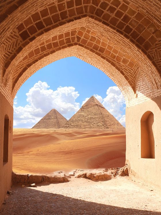 View through an arched doorway framing two pyramids on a sunny day, with a desert landscape and a bright blue sky with fluffy clouds.