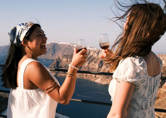 Two women laughing and holding wine glasses on a sunny cliffside, with a scenic view of the ocean and mountains in the background.