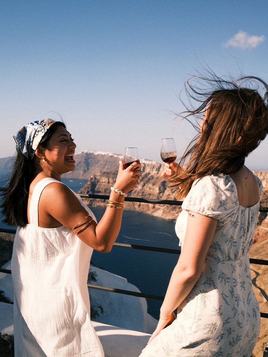 Two women laughing and holding wine glasses on a sunny cliffside, with a scenic view of the ocean and mountains in the background.