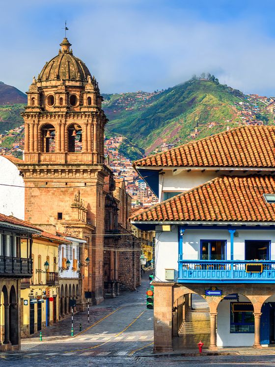 Historic street in Cusco, Peru, with colonial architecture; a bell tower rises against a backdrop of green hills under a blue sky.