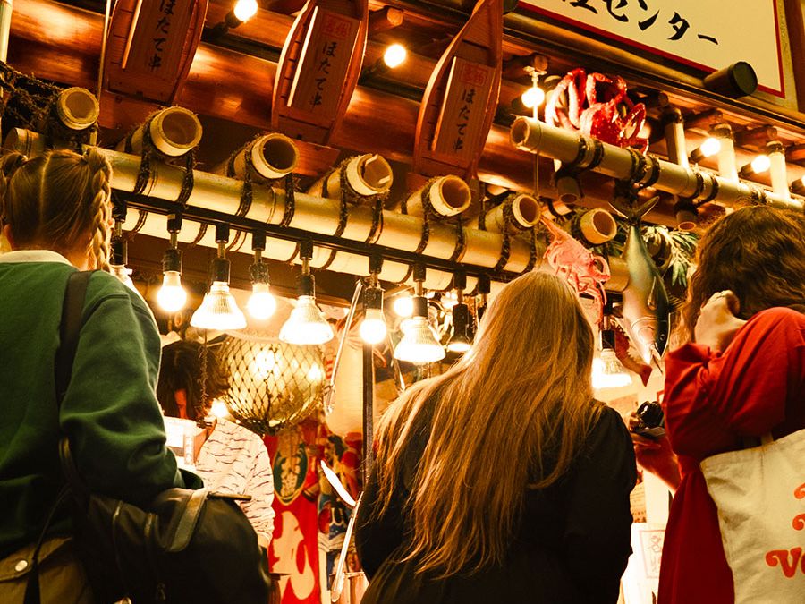 Three shoppers at a brightly lit Japanese seafood stall with hanging bulbs, wooden boat decorations and a large sign in Japanese.