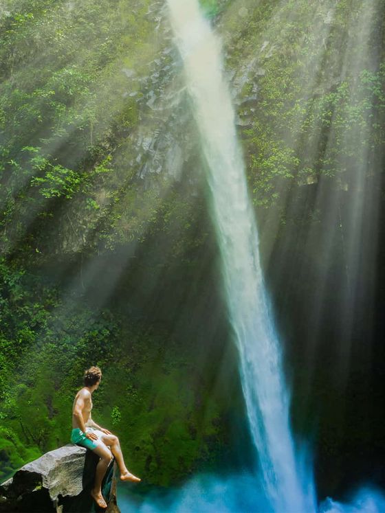 Person sitting on a rock, gazing at a tall waterfall surrounded by lush greenery, with sunlight streaming through the trees.