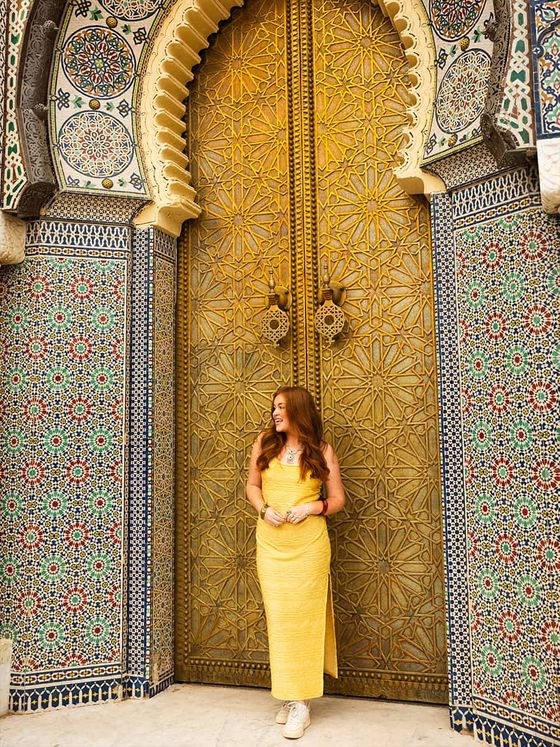 A woman in a yellow dress stands smiling in front of an ornate, gold-colored door with intricate mosaic tiles.