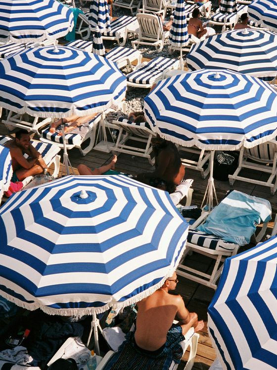 A busy beach scene with people relaxing under blue and white striped umbrellas on lounge chairs, creating a vibrant and sunny atmosphere.