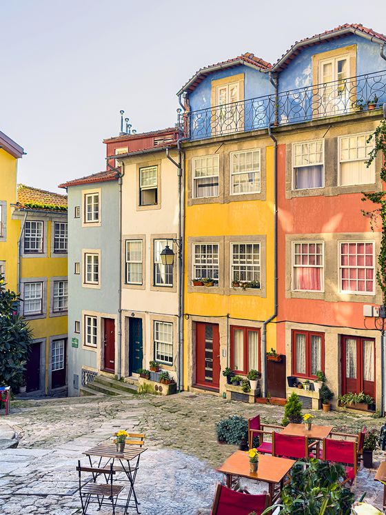 Colorful narrow European street with brightly painted houses, cobblestone path, small tables with red chairs, and vibrant potted plants.