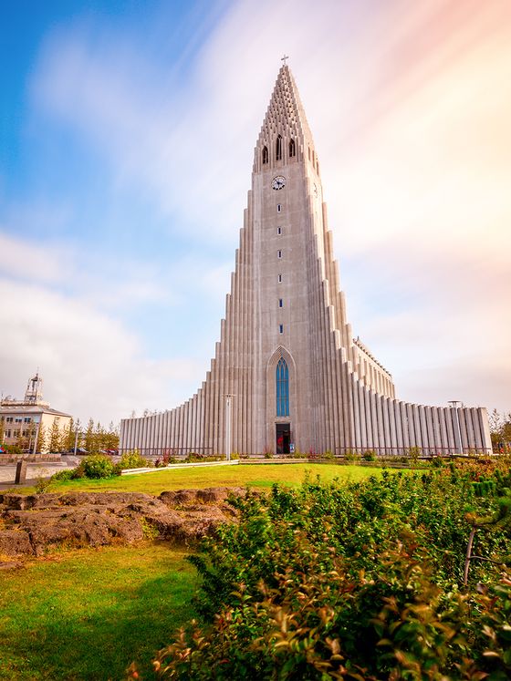Tall, modern church with a striking, stepped facade stands against a bright sky, surrounded by green grass and shrubs.