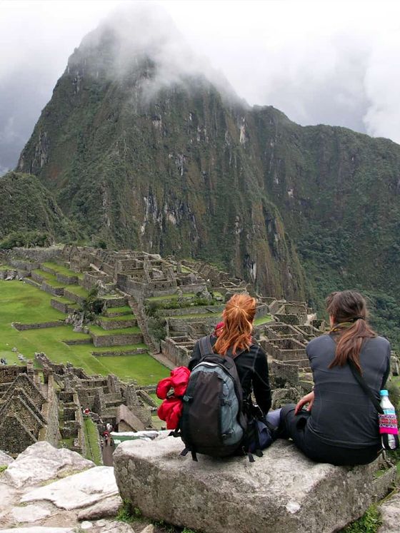Two people sit on a rock overlooking the ancient ruins of Machu Picchu, surrounded by lush greenery and misty mountains.