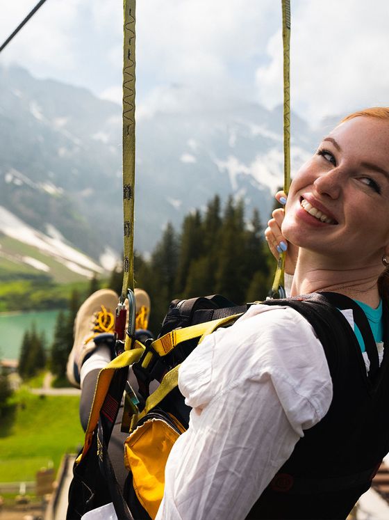 A woman smiles joyfully while zip-lining over a lush landscape with mountains and a lake in the background.