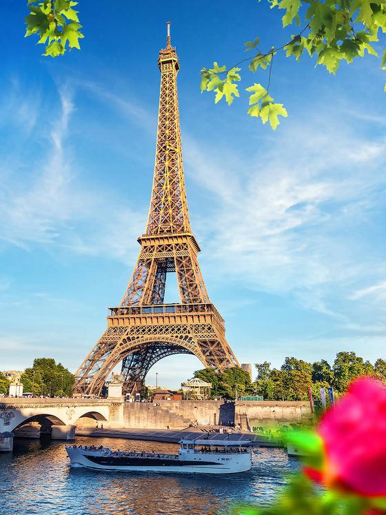 The Eiffel Tower with a boat on the Seine River, surrounded by green foliage and a blurred red flower in the foreground, under a blue sky.